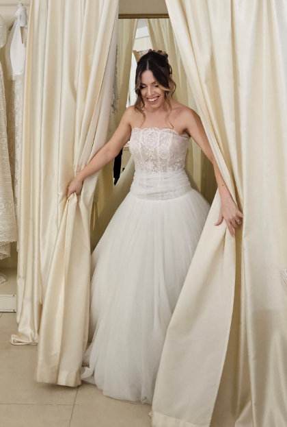 Sposa sorridente che esce dal camerino con un abito senza spalline, corpetto in pizzo e gonna ampia