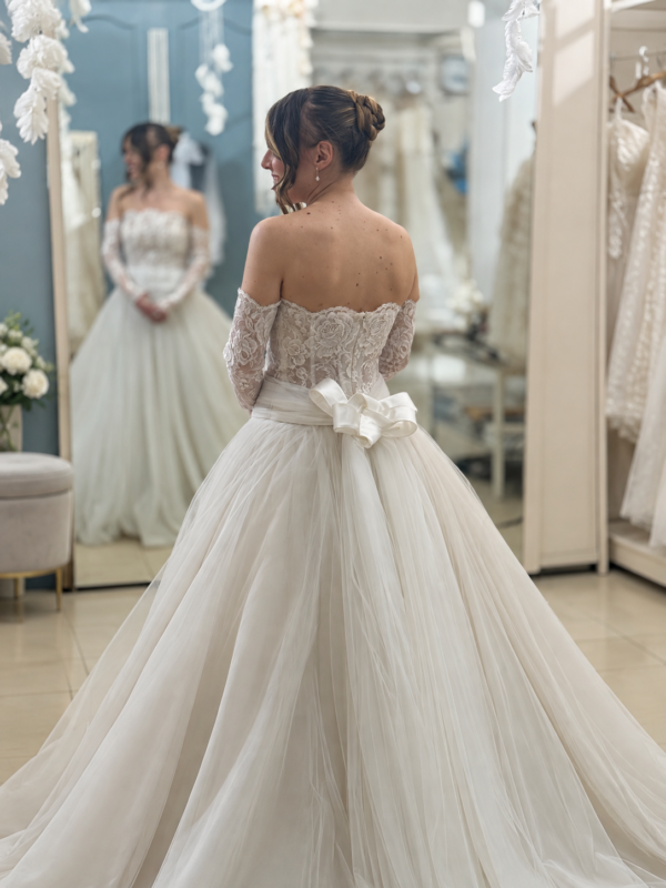 Prova davanti allo specchio di un abito da sposa con corpetto e maniche in pizzo, gonna in tulle e fiocco